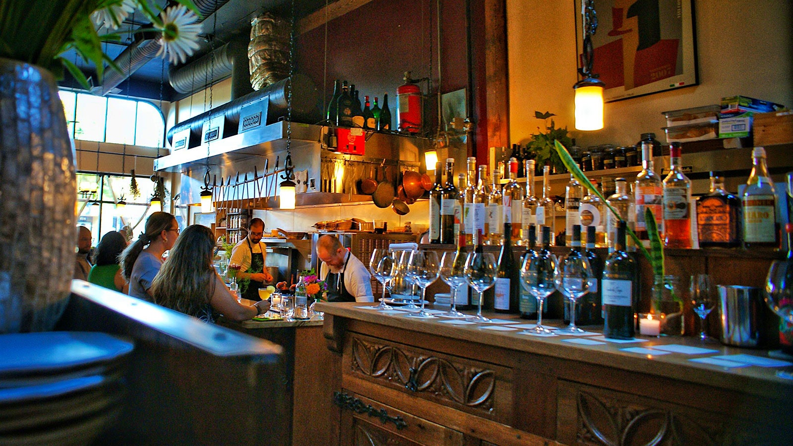 People in the dining room of Altura, with long wooden tables, many bottles of wine, wineglasses and an open kitchen